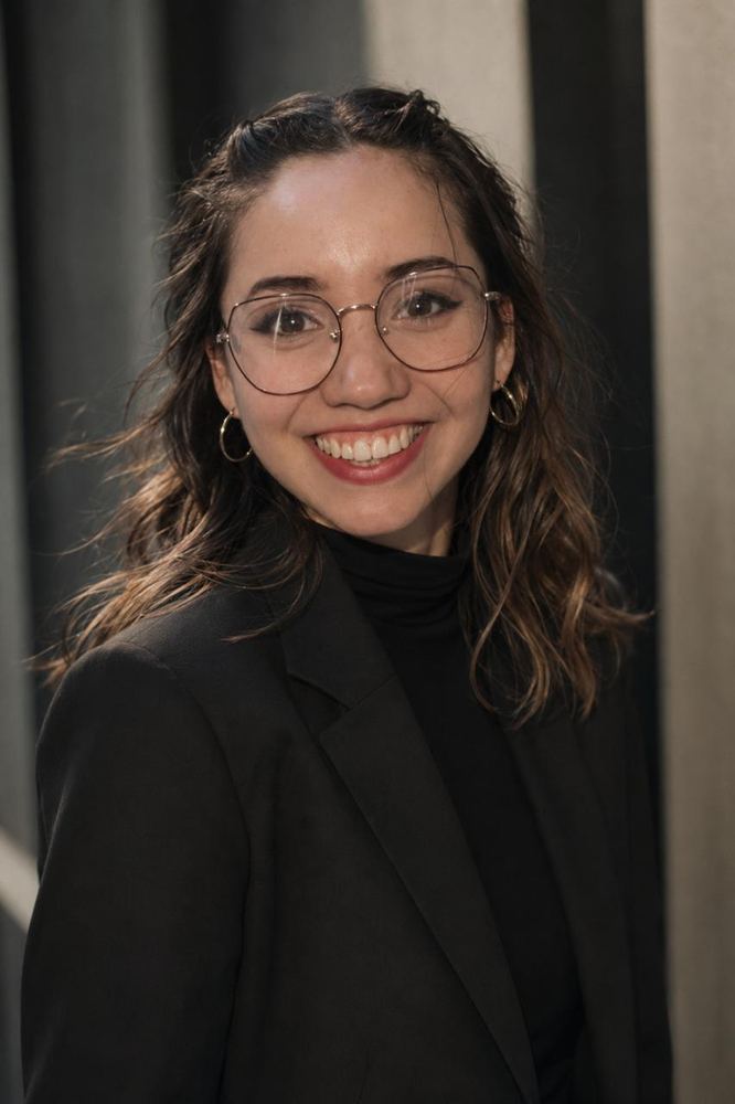 Erica in a black blazer and turtleneck, smiling, against a slatted wall