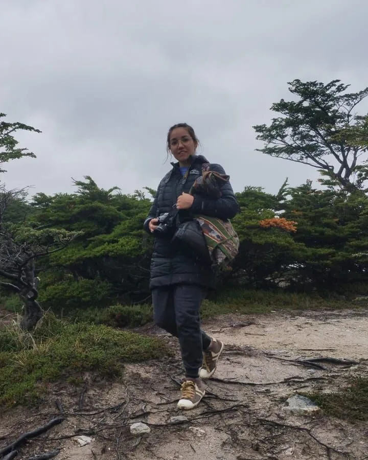 Erica on a dirt trail in a dark puffer jacket, a camera in her hands and a woven bag on her shoulder, low wind-bent trees and a grey sky behind her